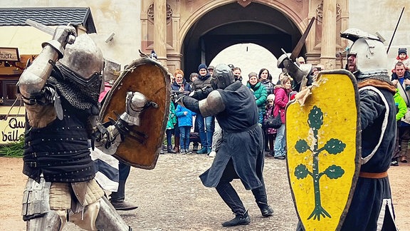Menschen in Ritterrüstung und mit Schild und Schwert in einem Schlosshof beim Kampf.