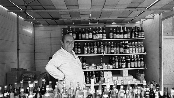 Schwarz-Weiß-Fotografie eines älteren Mannes in einem weißen Kittel, der in einem Raum voller alkoholischer Getränke steht