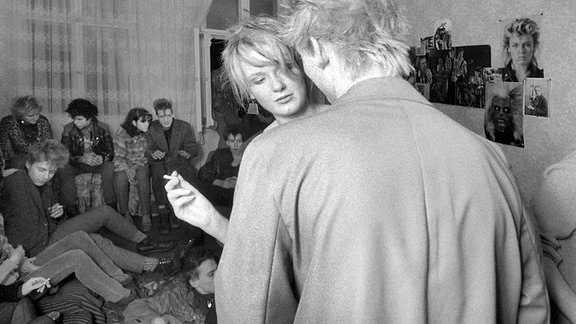 Schwarz-Weiß-Fotografie einer Partygesellschaft in einem Raum mit 80er-Jahre-Postern an den Wänden; im Vordergrund eine junge Frau mit Zigarette in der Hand, die einen jungen Mann anschaut 