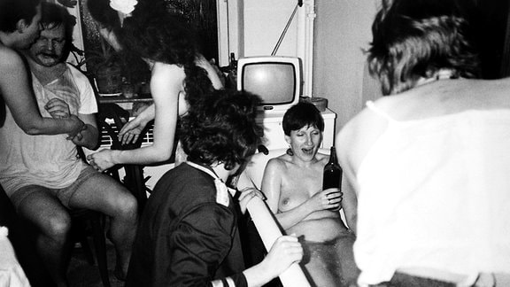 Schwarz-Weiß-Fotografie einer Gruppe Menschen, die zusammen feiert; im Zentrum des Bildes liegt eine lachende junge Frau nackt in einer Badewanne und hält ein Bier in der Hand