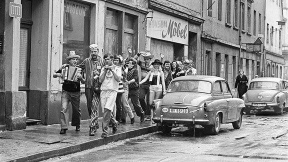 Schwarz-Weiß-Fotografie eine Faschingsgesellschaft, die in den 80er-Jahren durch eine Straße mit Geschäften und parkenden Autos zieht