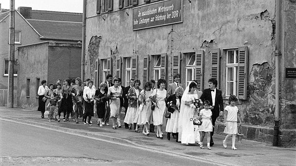 Schwarz-Weiß-Fotografie einer Hochzeitsgesellschaft, die an einem heruntergekommen Haus mit einem DDR-Banner vorbeizieht
