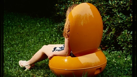 Rückseite eines Kunststoffstuhls in einem Garten, die Beine einer Frau sind zu sehen