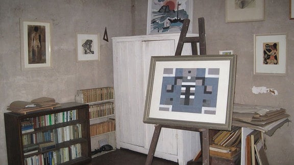 Blick in ein Zimmer: viele Bilder an der Wand, ein niedriger Bücherschrank, ein weißer Schrank und eine Bild auf einer Staffelei.
