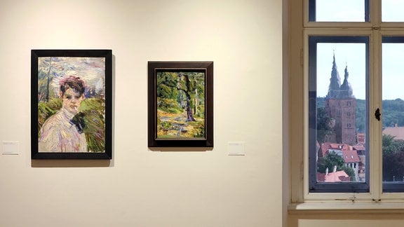 An einer hellen Wand hängen zwei Gemälde, auf der rechten Seite ein Fenster