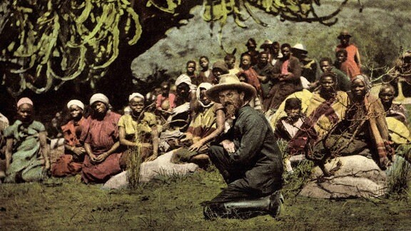 Ein weißer Mann mit Hut und Brille kniet auf dem Boden, hinter ihm sitzen Afrikaner