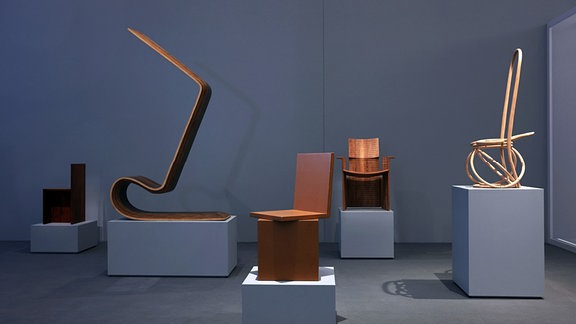  Ein Blick in die Ausstellung "A Chair and You" im GRASSI Museum, verschiedene Sitzmöglichkeiten, die alle aus Holz zu bestehen scheinen, sind in einem grauen Raum auf kleinen Podesten ausgestellt.