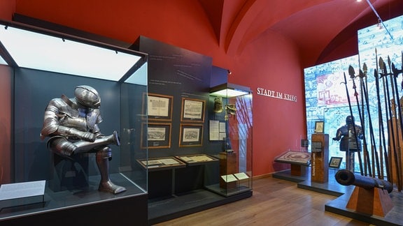Blick in eine Ausstellung mit Ritterüstungen, Speeren und historischen Stadtansichten.