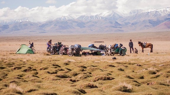 Ein Foto, eine Gruppe von Menschen mit Pferd, Motorrad und Zelt steht auf einer Wiese, im Hintergrund schneebedeckte Berge.