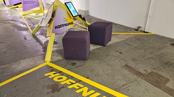 Zwei Sitzhocker in einer Ausstellung laden zum Hinsetzen ein, auf dem Boden steht in gelber Schrift das Wort "Hoffnung".