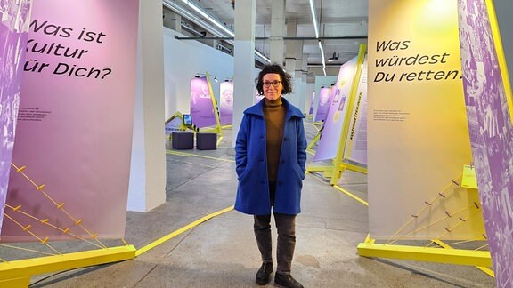 Eine Frau in einem blauen Mantel steht zwischen zwei Aufsstellern in einer Ausstellung. Kulturretter:innen 