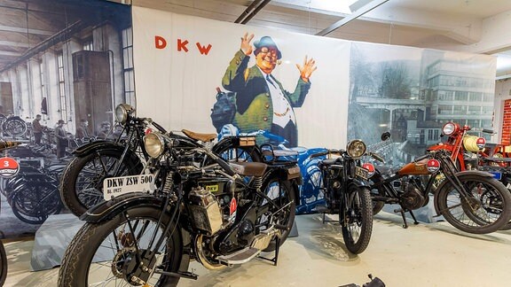 In einer Halle in einem Museum stehen mehrere historische Motorräder. 