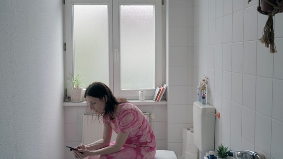 Eine Frau sitzt in einem rosa Kleid auf der Toilette und schaut aufs Handy. 