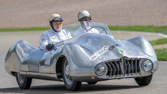 Zwei Männer mit Helmen sitzen in einem kleinen silberfarbenen Sportwagen.