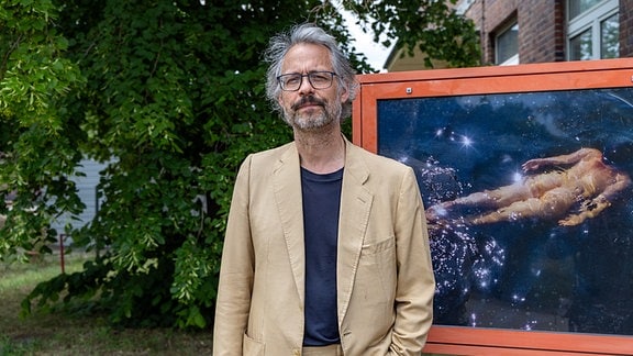 Festivalleiter Aljoscha Begrich, ein Mann mit mittellangem grauem Haar, Bart und beigem Jackett, steht vor einem Plakat, auf dem eine nackte schwimmende Person zu sehen ist.