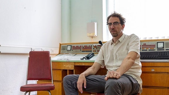 Festivalmacher Christian Tschirner,  ein mann mit kurzen Haaren, kurzem Bart und Brille mit weißem Hemd sitzt auf einem Stuhl. Im Hintergrund ist ein DDR-Schaltpult zu sehen.