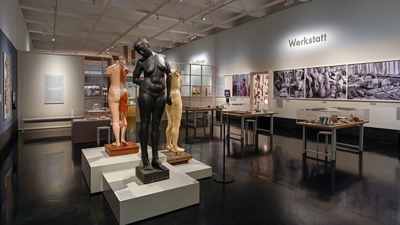 Blick in eine Ausstellung, auf der linken Seite stehen Statuen, auf der rechten Seite hängen schwarz-weiß Bilder an den Wänden