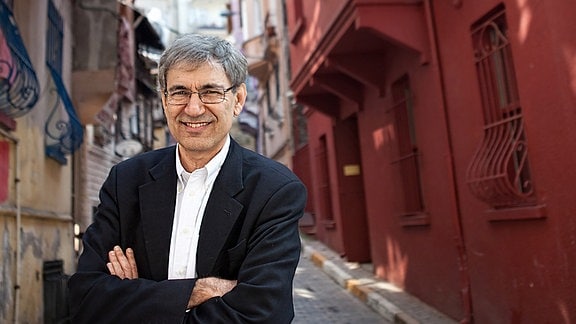 Orhan Pamuk, ein Mann mit grauen Haaren und einer Brille, steht mit verschränkten Armen auf einer Straße. 