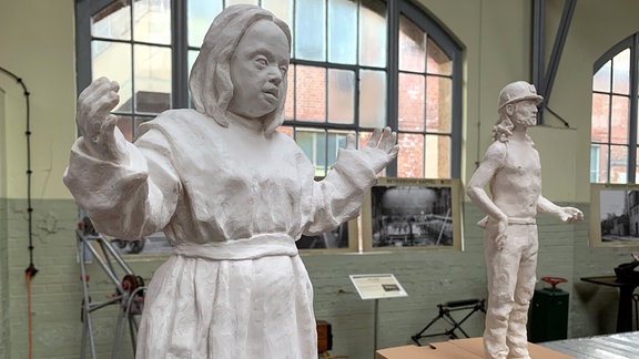 Skulptur eines Engels mit ausgebreiteten Armen und Zügen eines Menschen mit Down-Syndrom; im Hintergrund ein Bergmann im Stil eines Wismut-Kumpels