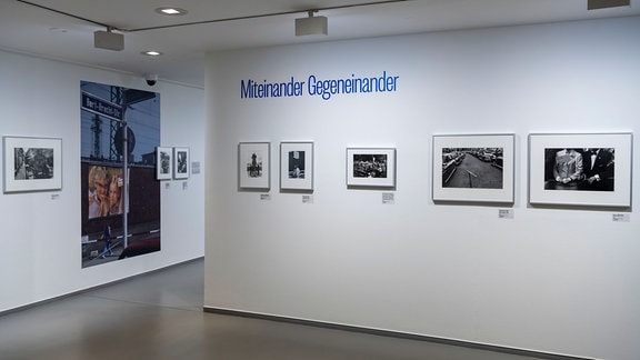 Zu sehen ist ein Blick in die Ausstellung, schwarz-weiß Fotografien hängen an der Wand, in Blauer Schrift steht "Miteinander Gegeneinander" an der Wand