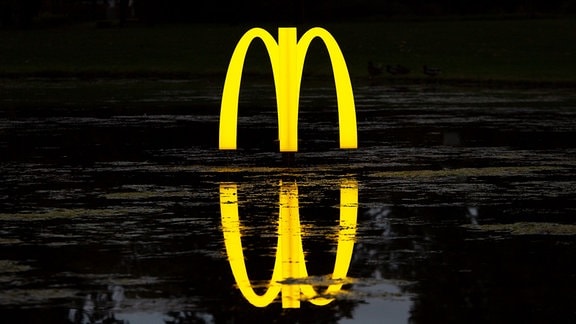 Das gelb leuchtende McDonald's-M erhebt sich auf einem nächtlichen Gewässer als Springbrunnen. 