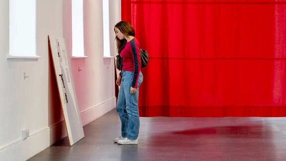 Ausstellungsansicht "Sarah Pierce: Scene of the Myth" in der Galerie für Zeitgenössische Kunst Leipzig, ein roter Vorhang hängt im Hintergrund, vorne schaut sich eine Frau ein Kunstwerk an.