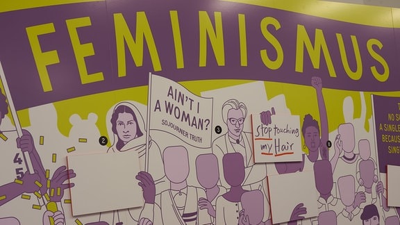 Ein an die Wand gemaltes Bild im Comic-Style, auf dem "Feminismus" in Großbuchstabensteht, Frauen halten verschiedene Transparente