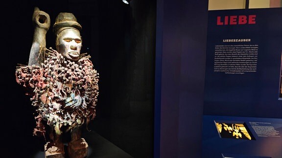 Eine mit Nägeln und Klingen beschlagene Figur steht in einer beleuchteten Vitrine in einem Ausstellungsraum.