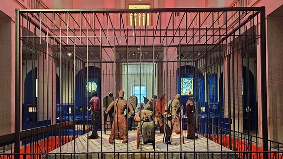 Ein Käfig mit menschengroßen Puppen steht in einem Ausstellungsraum.