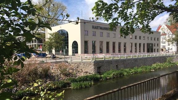 Ein lang gestrecktes, helles Gebäude mit hohen Fenstern und der Aufschrift "Hartmann Fabrik" steht an einem Fluss, von grünen Ästen umrankt.