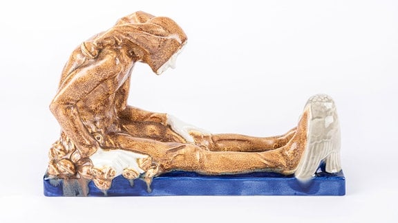 Figur "Till Eulenspiegel" aus sandfarbenem Porzellan sitzt vorn übergebeugt auf einem blauen Sockel.