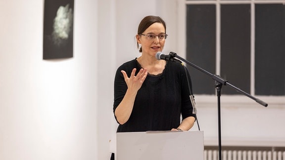 Jule Reuter, eine Frau mit schwarzer Kleidung und Brille steht an einem Rednerpult.