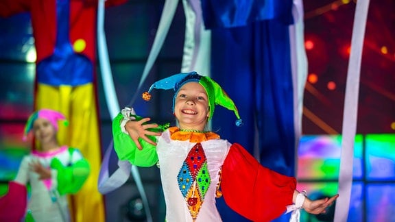 Ein kleines Mädchen tanzt in einem bunten Clowns-Kostüm auf einer Bühne.