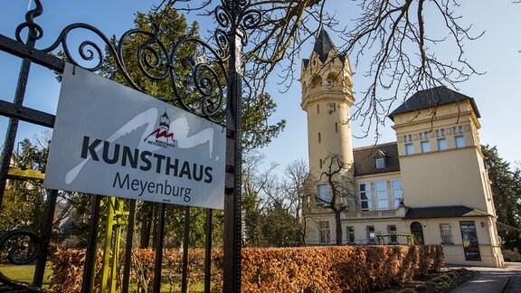 Kunsthaus Meyenburg: ein burgenähnlicher Bau mit heller Fassade und Turm in einem Park, davor ein geöffnetes Metalltor