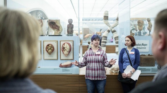 Zwei junge Frauen stehen in einem Museum und sprechen zu einer Besuchergruppe.