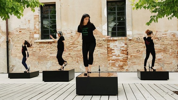 Videostill einer Performance: Auf schwarzen Podesten stehen Frauen in schwarzer Kleidung mit Kopfhörern und tanzen.