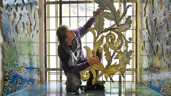 Ein Mann sitzt vor einem bodentiefen Kirchfenster und hält sein Kunstwerk aus Emaille in der Hand - es zeigt in gold Blätter und einen Vogel an einem Ast.