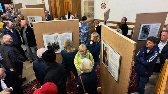 Ausstellungsbesucher schauen sich in einer Kirche aufgestellte Tafeln an, auf denen Fotos und Text zu sehen sind.