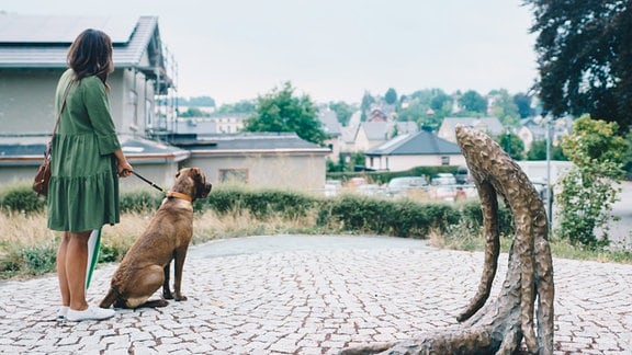 Eine Frau mit Hund steht neben einer Bronze-Skulptur in Form eines geknickten Zweiges.