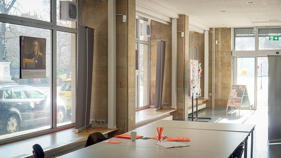 Ein Konferenztisch steht in einem Innenraum. An den bodentiefen Fenstern, die zur Straße zeigen, hängen Fotografien.