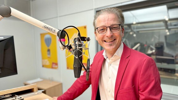 Ein Mann in rotem Anzug, weißem Hemd und Brille steht in einem Studio vor einem Mikrofon und lächelt in die Kamera.