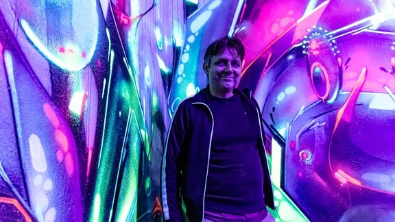 Ein lächelnder Mann in Traningsjacke vor einer grellbunten, violett-blauen Wand: Martin Gerth aka Noise, Künstler aus Magdeburg