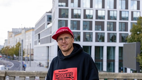 Ein Mann mit roter Basecap und Brille steht lächelnd auf einer Brücke modernen Blocks: Nils Klebe von den Da Rookies