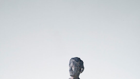 Die graue Skulptur eines Mannes in schlichter Kleidung, der den Kopf nach rechts dreht.