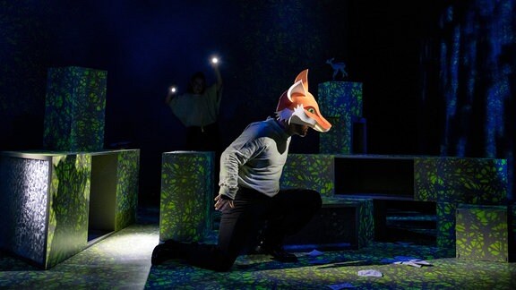 Ein Mensch mit Fuchsmaske läuft über die Dresdner Bühne, im Hintergrund stehen grüne Quader im Halbdunkel.