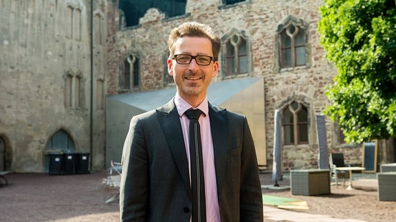 Ein Mann mit Anzug, kurzem Haar und Brille steht vor einem historischen Gebäude und lächelt in die Kamera