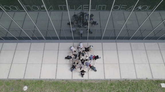 Blick von oben auf eine Gruppe in einer Kreisformation vor einer Glasfassade.