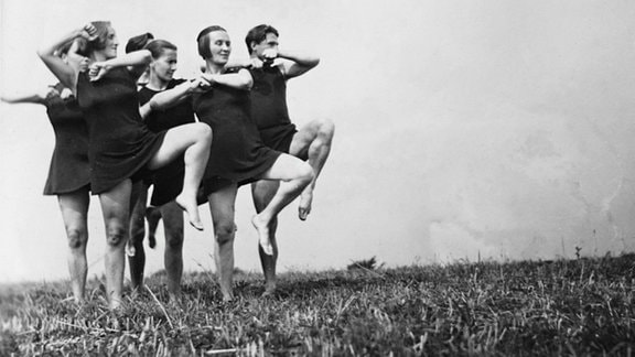 Schwarz-Weiß-Bild von einer Personengruppe, die auf tänzerisch auf einem Bein auf einer Wiese stehen und das andere Bein angewinkelt heben.