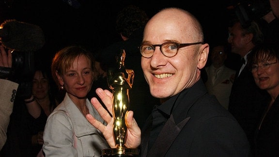 Ulrich Mühe trägt einen schwarzen Anzug und hält stolz den Filmpreis Oscar in die Kamera.