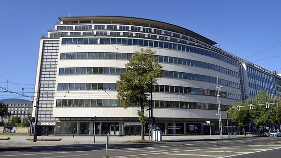 Ein modernes längliches mehrstöckiges Gebäude mit heller Fassade vor blauem Himmel. 